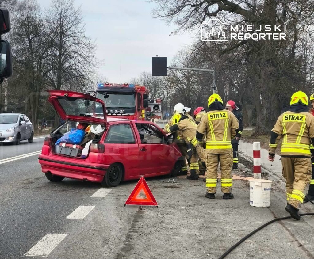 Grupa strażaków w żółtych kombinezonach udziela pomocy poszkodowanemu w czerwoną osobówkę na poboczu drogi.