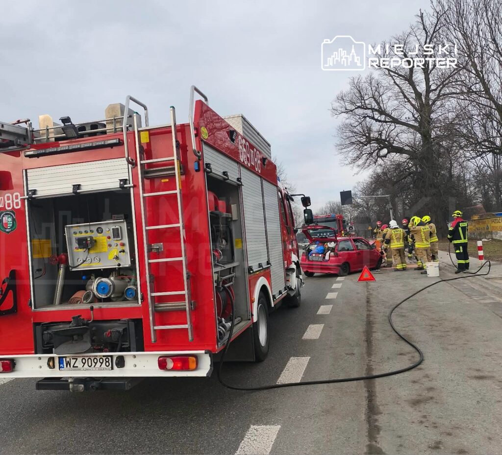 Zespół ratunkowy pracuje przy czerwonym samochodzie osobowym na poboczu drogi, obok wozu strażackiego.