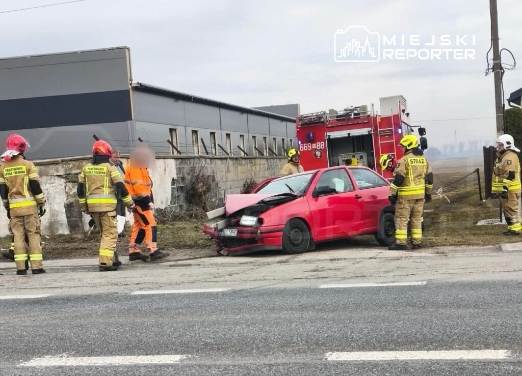 Grupa strażaków w mundurach i kaskach ocenia uszkodzony czerwony samochód osobowy przy drodze, obok wozu strażackiego.