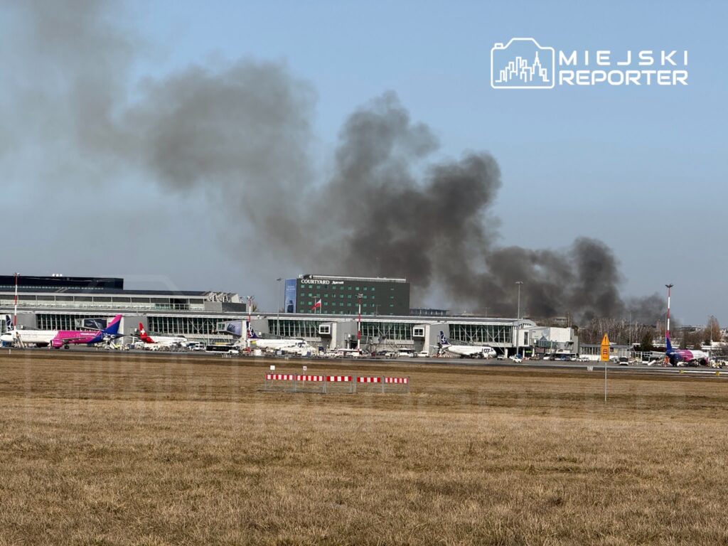 Czarne chmury dymu unoszą się nad nowoczesnym terminalem lotniska, w tle widoczne samoloty na płycie lotniska.