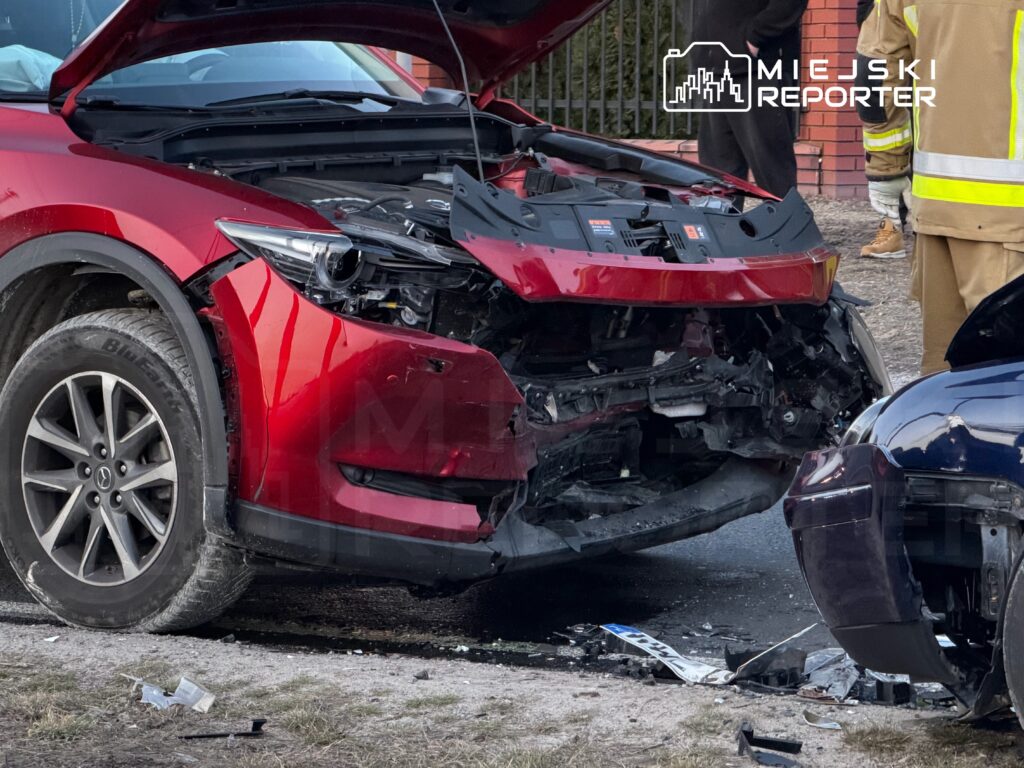 Czerwony samochód osobowy z uszkodzoną przednią częścią stoi na drodze, obok niego widać strażaków i gapiów.