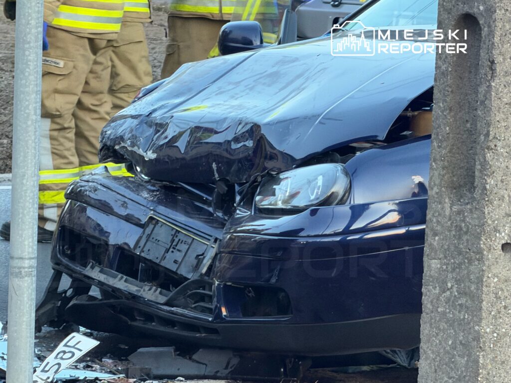 Zniszczony niebieski samochód osobowy z wgnieceniem na przodzie stoi przy słupie, obok strażacy w mundurach.