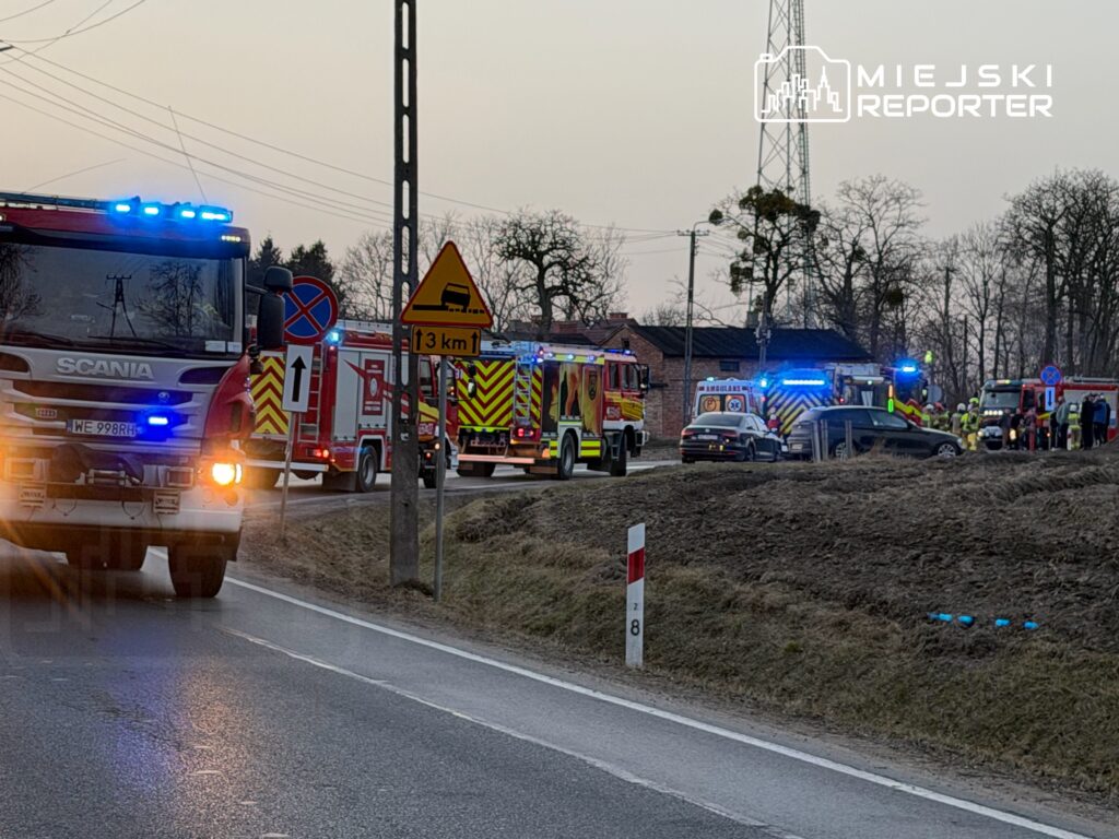 Zespół ratunkowy z wozami strażackimi i karetką pracuje przy drodze, obok znaku drogowym i budynku.