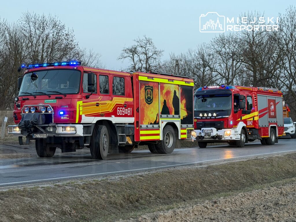 Dwa wozu strażackie z włączonymi sygnałami świetlnymi stoją na poboczu drogi w otoczeniu drzew.