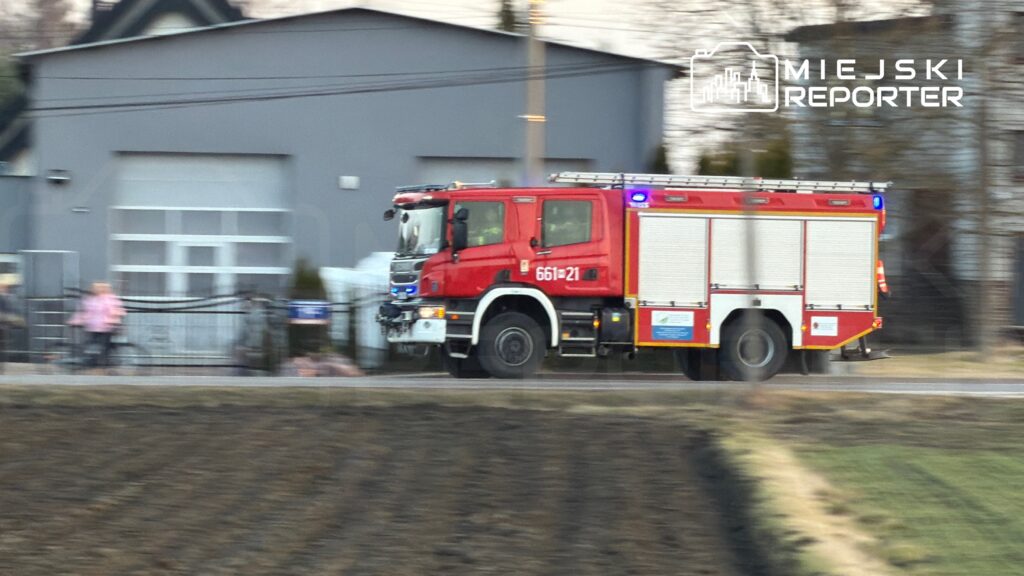 Czerwony wóz strażacki z włączonymi sygnałami świetlnymi przejeżdża obok budynku mieszkalnego.