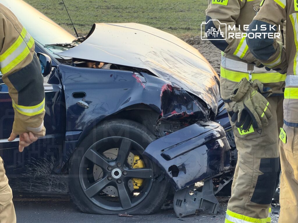 Pojazd osobowy z uszkodzoną przednią częścią, obok stoją strażacy w mundurach roboczych.