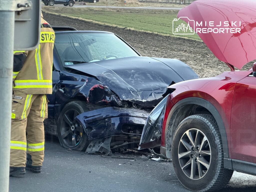 Strażak w mundurze z odblaskowymi paskami ocenia uszkodzone pojazdy po zderzeniu na drodze.