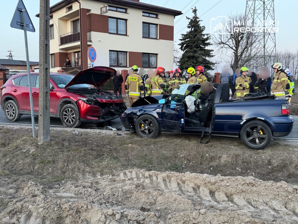 Czerwony SUV z uszkodzonym przodem stoi obok niebieskiego kabrioletu, przy którym pracują strażacy.