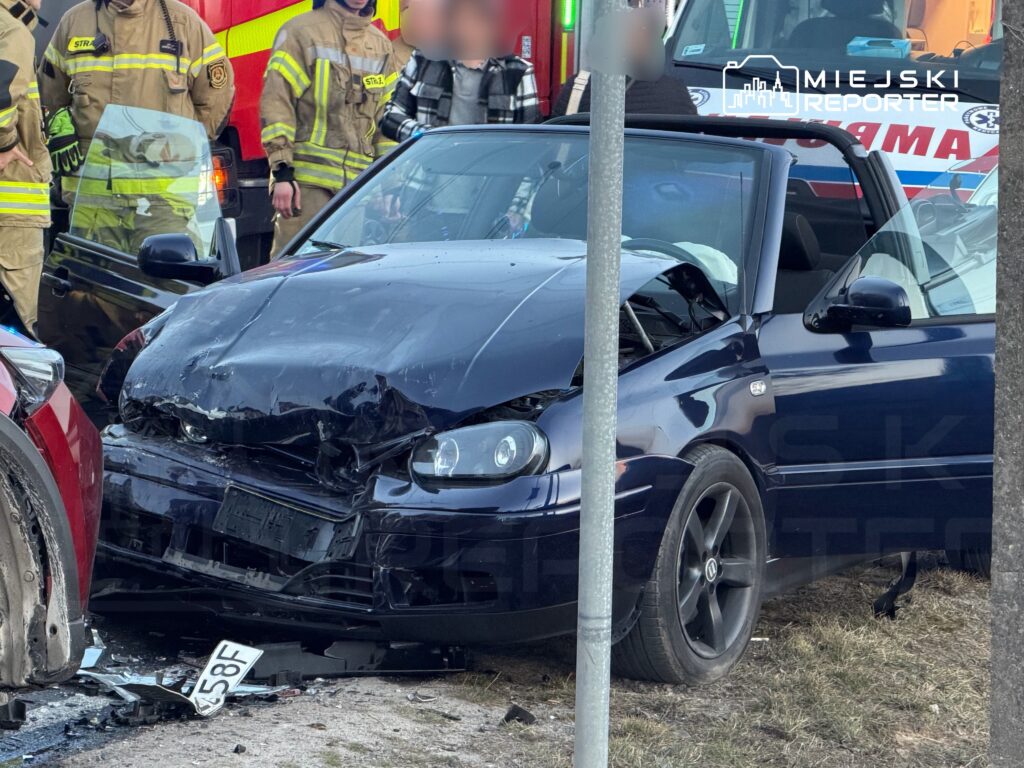 Czarny samochód osobowy z uszkodzoną przednią częścią zaparkowany obok czerwonego pojazdu, wokół stoją strażacy.