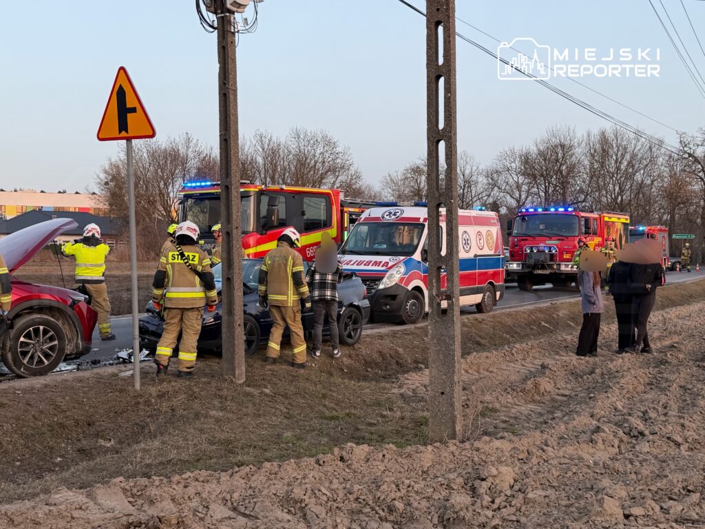 Grupa strażaków udziela pomocy przy wypadku drogowym obok uszkodzonych samochodów, w tle widać karetkę i pojazdy ratunkowe.
