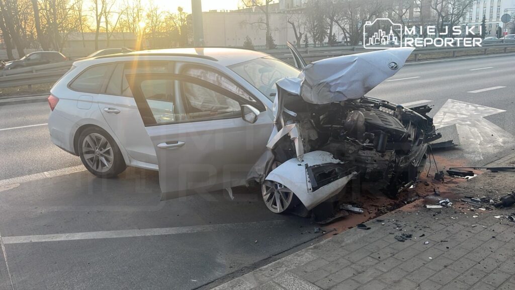 Uszkodzony samochód osobowy leży na poboczu drogi, z wyraźnymi uszkodzeniami przodu, obok znaku kierunkowego.