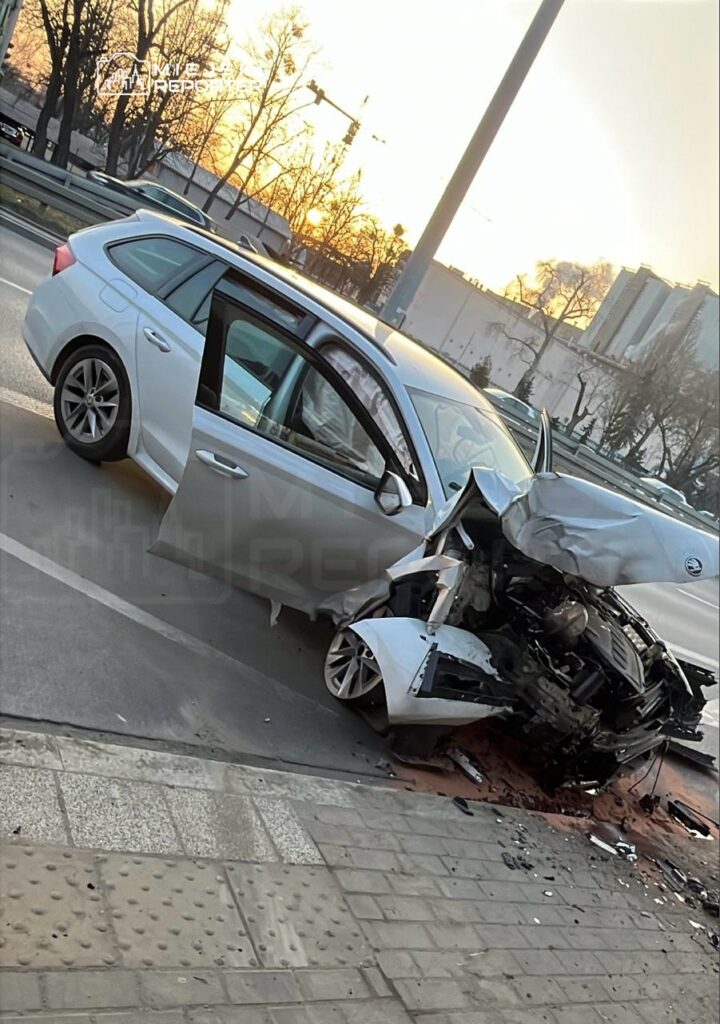 Uszkodzony samochód osobowy leży na poboczu drogi, z widocznymi uszkodzeniami przodu, obok otwartych drzwi.