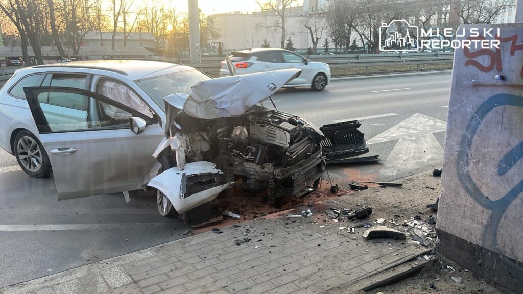 Zniszczony samochód osobowy leży na chodniku po zderzeniu, obok widoczne fragmenty karoserii i olej.