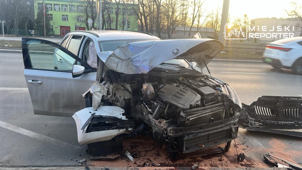 Rozbity samochód osobowy z uszkodzoną przednią częścią stoi na poboczu drogi, obok otwartych drzwi.