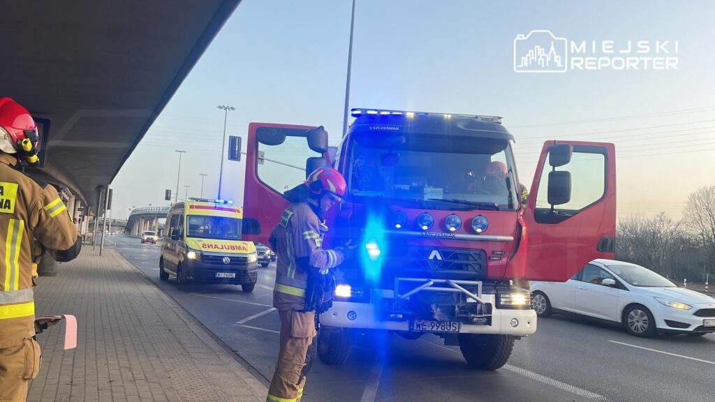 Strażak w hełmie kontroluje sprzęt przed wozem strażackim, obok przejeżdża karetka pogotowia.