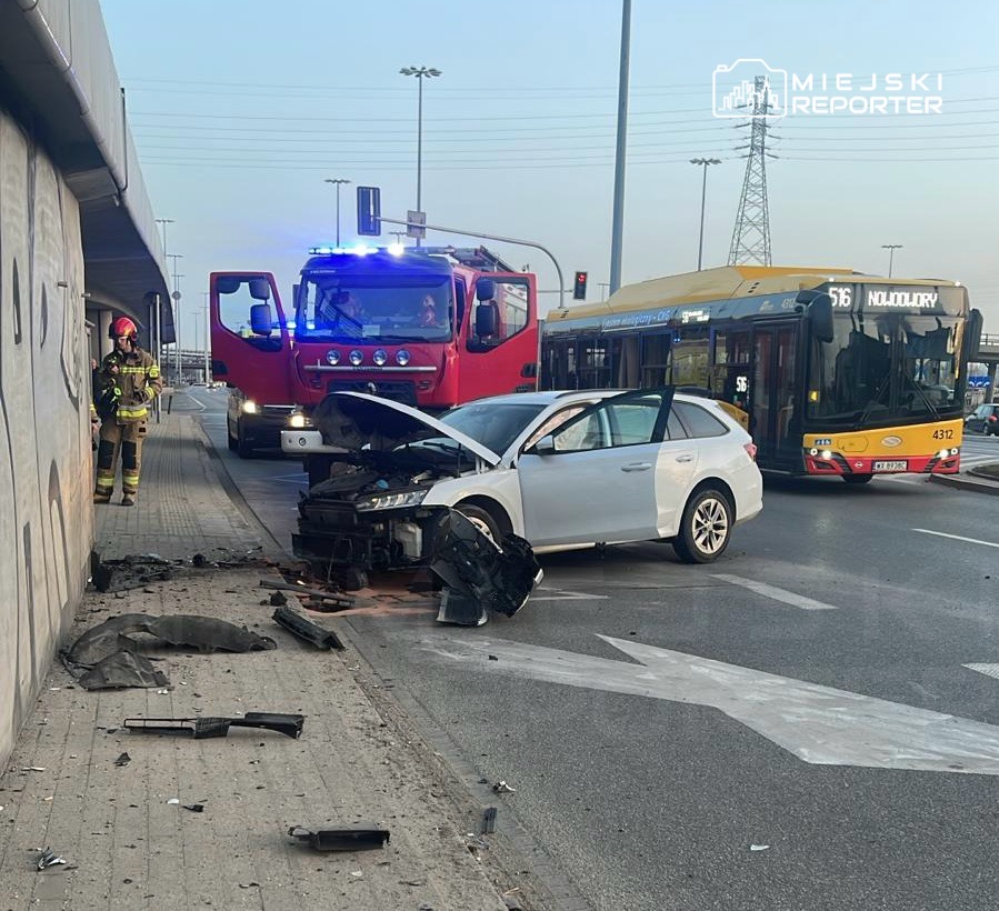 Wypadek drogowy z uszkodzonym samochodem osobowym, obok straż pożarna i autobus miejski na ruchliwej ulicy.