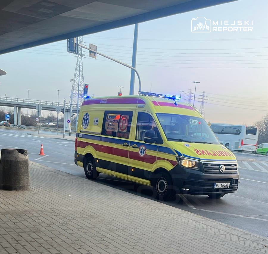Karetka pogotowia z włączonymi sygnałami świetlnymi stoi na poboczu drogi obok przystanku autobusowego.