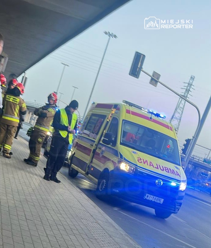 Zespół ratowników w mundurach stoi obok karetki pogotowia na chodniku, w tle widoczne sygnalizatory świetlne.