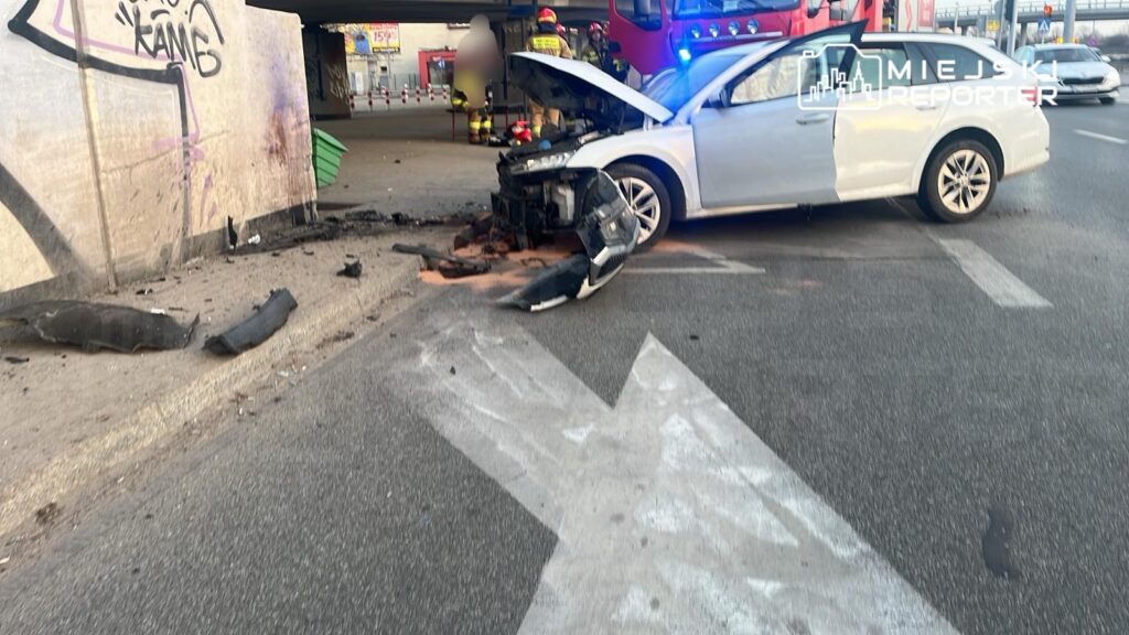 Zniszczony samochód osobowy z otwartą maską stoi na jezdni obok graffiti na ścianie, w tle widać strażaków.