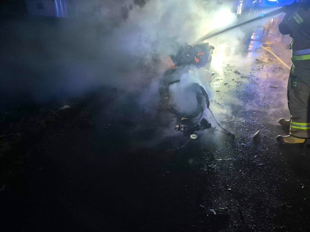 Strażak gaszący płonący wrak pojazdu na drodze, otoczony gęstym dymem i niebieskimi światłami.