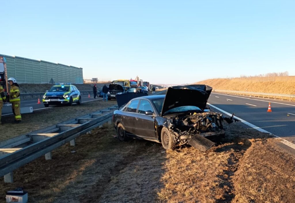 Czarny samochód osobowy z uszkodzoną przednią częścią stoi na poboczu drogi, obok pracują strażacy i służby drogowe.