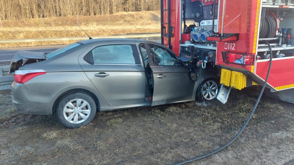 Zniszczony samochód osobowy stoi obok wozu strażackiego na poboczu drogi, z otwartymi drzwiami.
