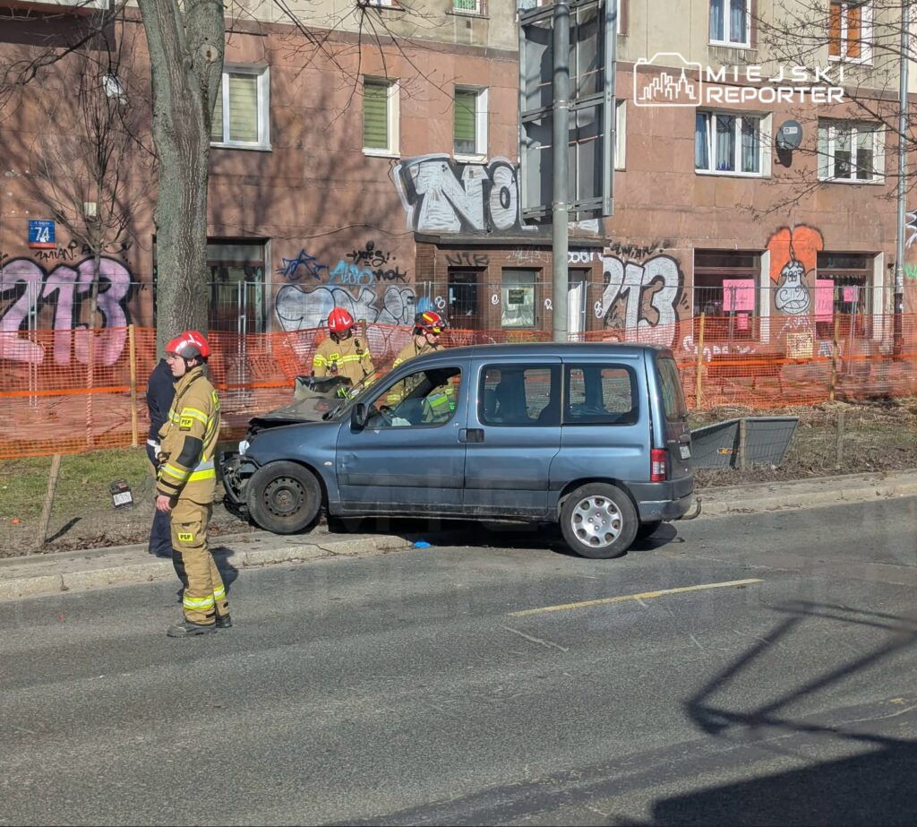 Strażacy pracują przy uszkodzonym niebieskim samochodzie osobowym zaparkowanym obok budynku z graffiti.