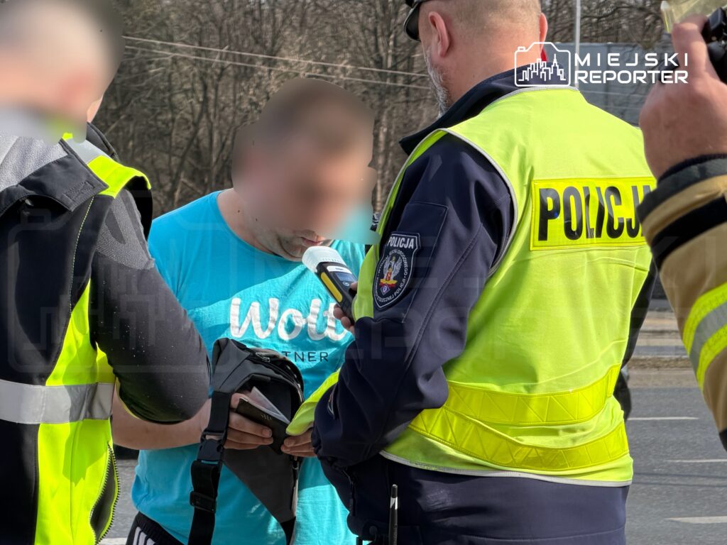 Mężczyzna w niebieskiej koszulce rozmawia z policjantem w kamizelce odblaskowej na drodze.