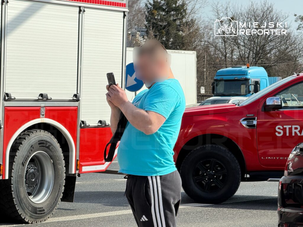 Mężczyzna w niebieskiej koszulce korzysta z telefonu w pobliżu wozu strażackiego na ruchliwej ulicy.