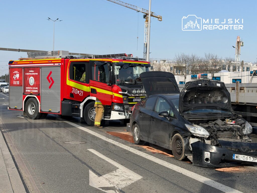 Straż pożarna z włączonymi sygnałami stoi obok uszkodzonego czarnego samochodu na ruchliwej ulicy.