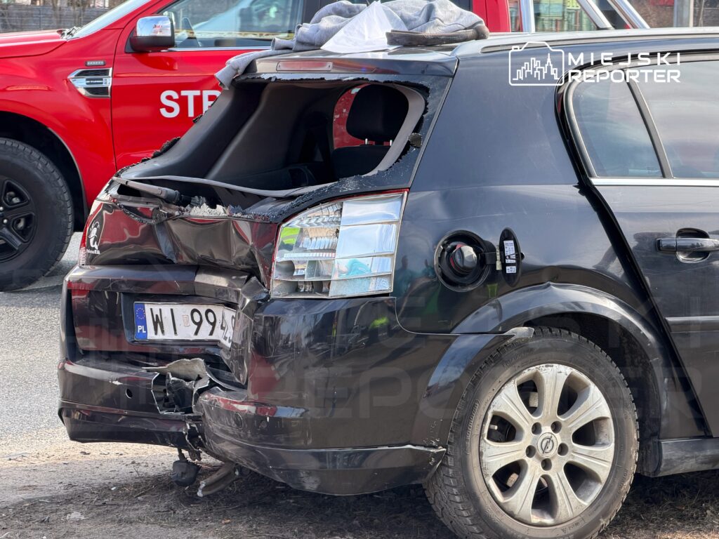 Uszkodzony czarny samochód osobowy z rozbitym tyłem stoi na poboczu drogi, w tle widoczny czerwony pojazd straży pożarnej.