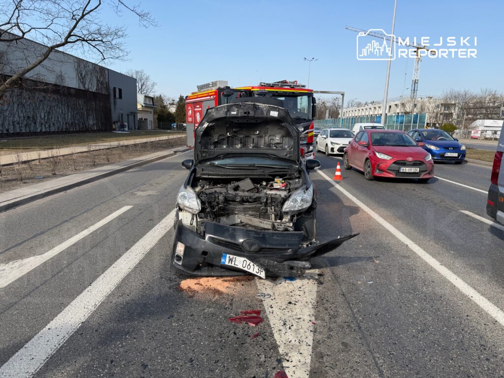 Uszkodzony samochód osobowy z otwartą maską na drodze, obok stoją pojazdy w korku, a w tle widoczny wóz strażacki.