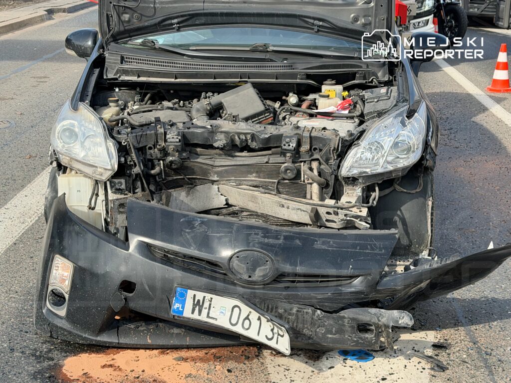 Zniszczony samochód osobowy z otwartą maską na jezdni, z widocznymi uszkodzeniami przodu i silnika.