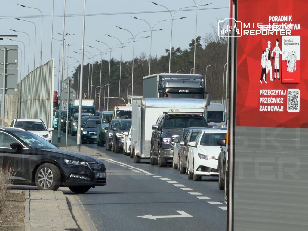 Kierowcy stoją w korku na drodze, obok widoczny billboard z napisem "Poradnik bezpieczeństwa. Przeczytaj, przeciwwcz zachowaj!"