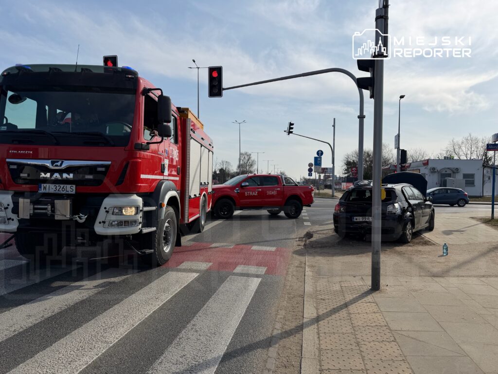 Czerwony wóz strażacki zatrzymany na skrzyżowaniu obok uszkodzonego czarnego samochodu, w tle sygnalizacja świetlna.