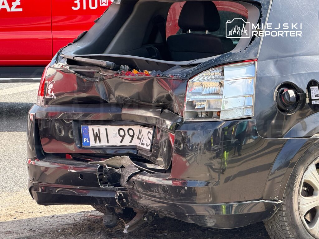 Czarny samochód osobowy z uszkodzoną tylną częścią zaparkowany na drodze, w tle czerwony pojazd służb ratunkowych.