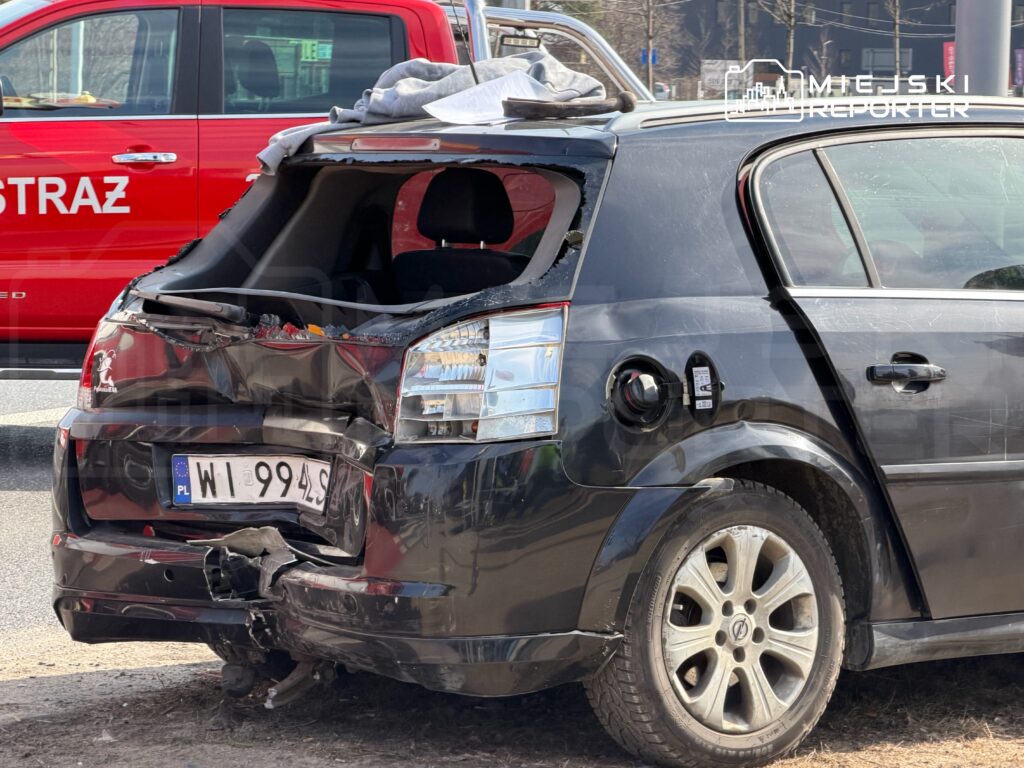 Uszkodzony czarny samochód osobowy z wgniecionym tyłem, zaparkowany obok czerwonego pojazdu straży pożarnej.