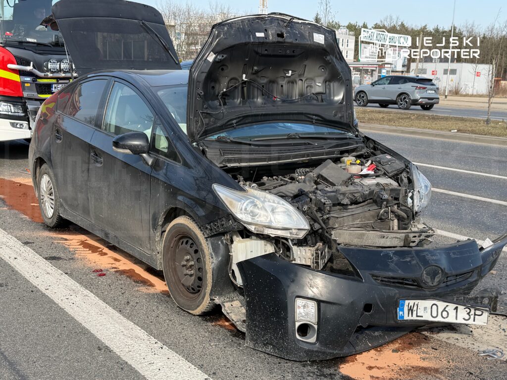 Czarny samochód osobowy z otwartą maską stoi na drodze, obok widoczne ślady płynów eksploatacyjnych.