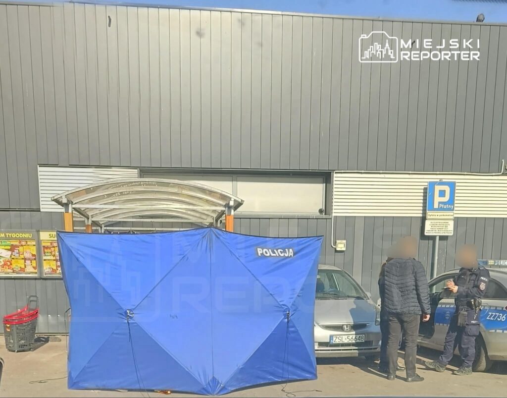 Policjanci rozmawiają z dwoma osobami przy niebieskim namiocie oznaczonym "POLICJA" przed budynkiem handlowym.