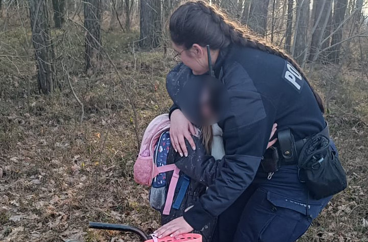 Policjantka przytulająca dziecko z plecakiem w lesie, obok stoi różowy rowerek.