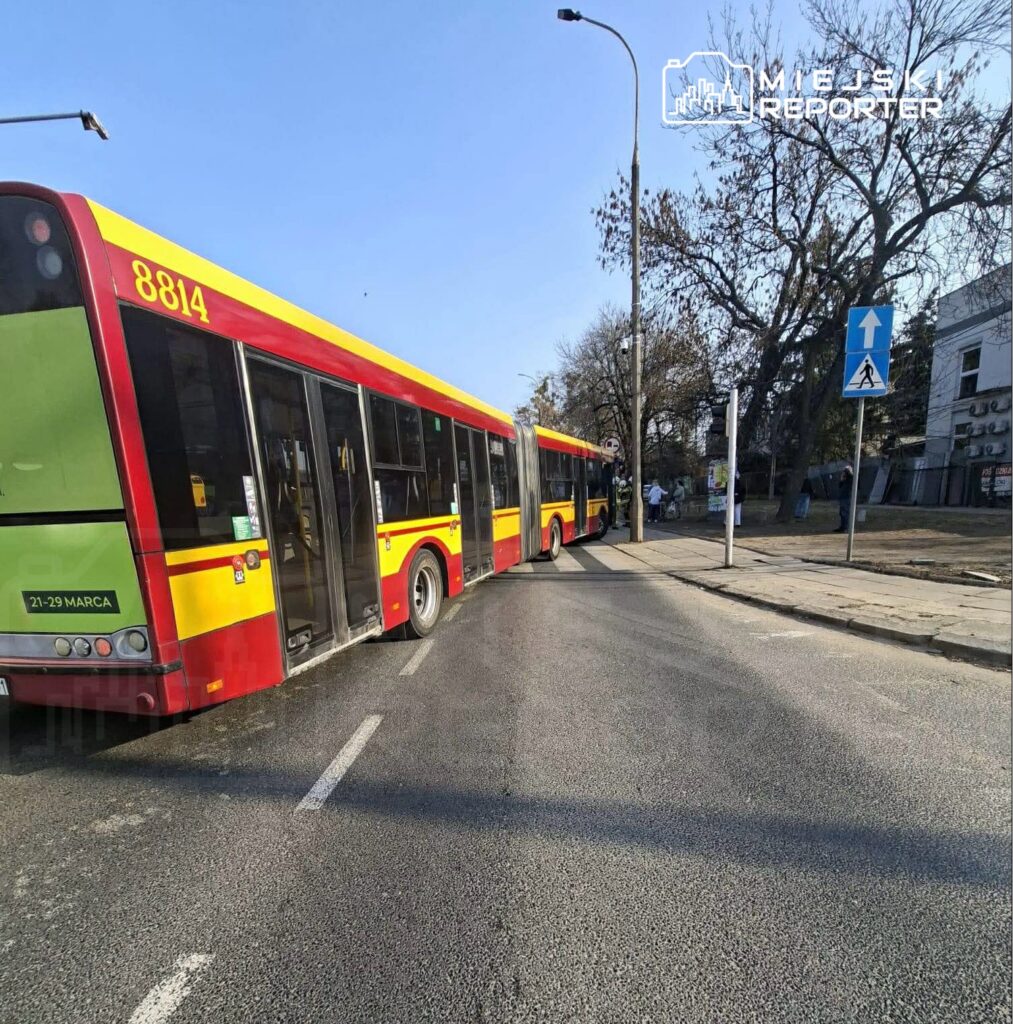 Autobus miejski skręcający w prawo na ulicy, obok znaku drogowym i drzewami w tle.