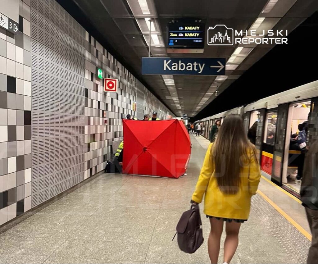 Kobieta w żółtym płaszczu idzie w stronę peronu metra, obok czerwony namiot i pasażerowie wchodzący do pociągu.