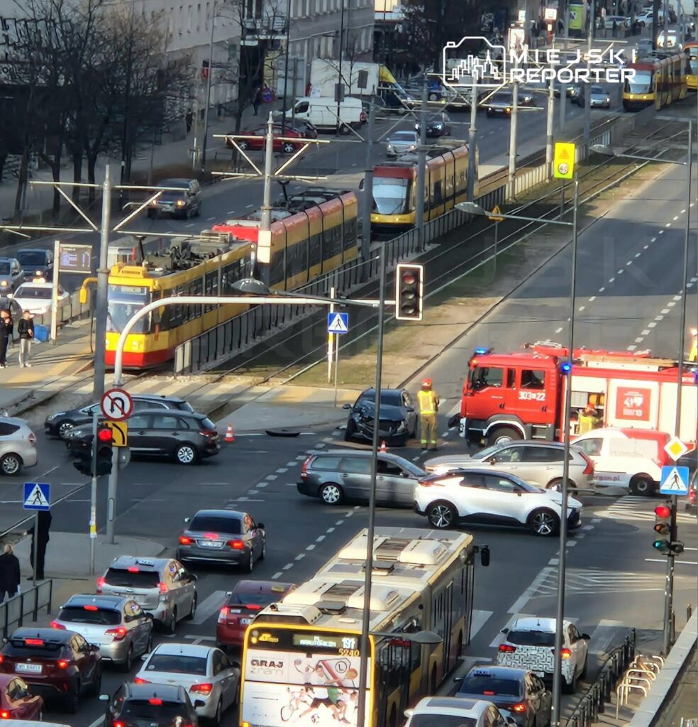 Na skrzyżowaniu ulicy widać zderzenie dwóch samochodów, obok stoi wóz strażacki z włączonymi sygnałami.