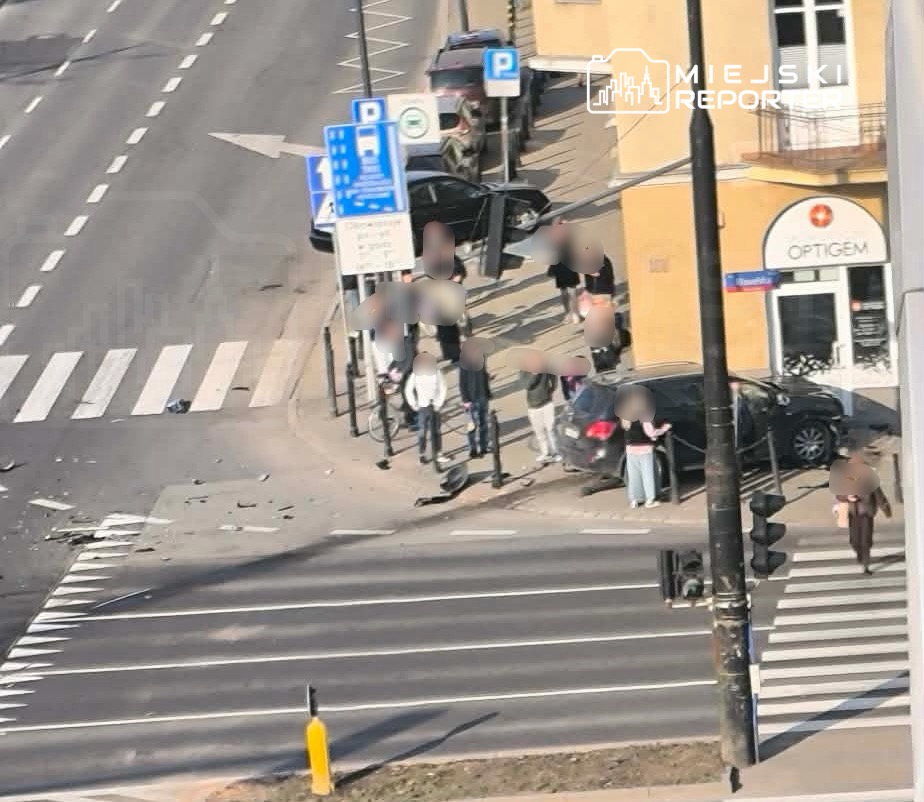 Grupa osób gromadzi się na chodniku przy ulicy, obok uszkodzonego samochodu, w tle widoczne znaki drogowe i budynek.