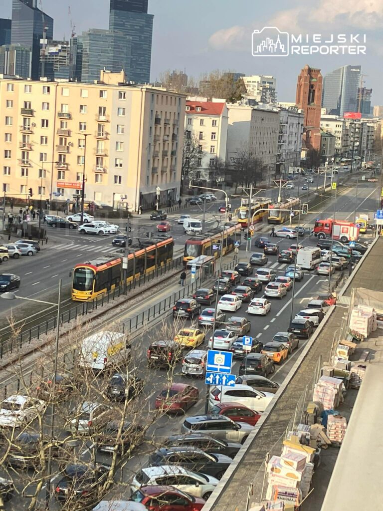 Tramwaj przejeżdża obok zatłoczonej ulicy w mieście, w tle nowoczesne wieżowce i budynki mieszkalne.