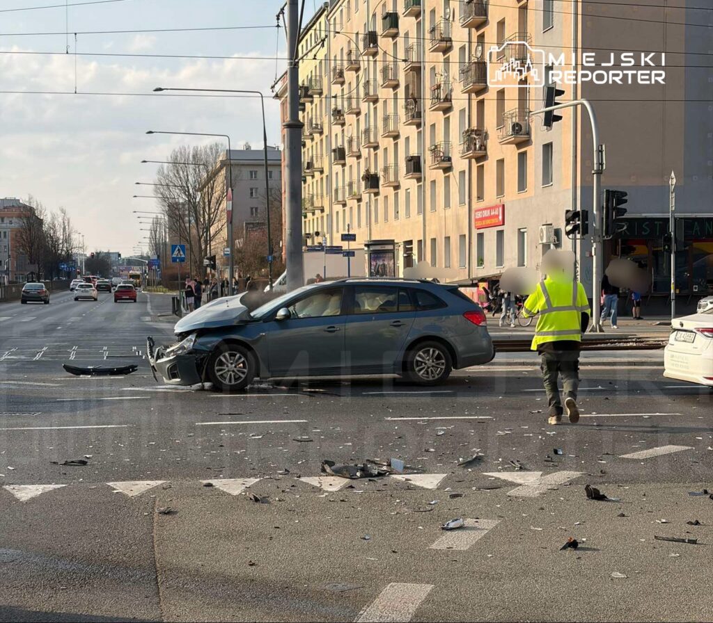 Uszkodzony samochód osobowy stoi na skrzyżowaniu, obok biegnący mężczyzna w żółtej kamizelce odblaskowej. Wokół leżą fragmenty pojazdu.