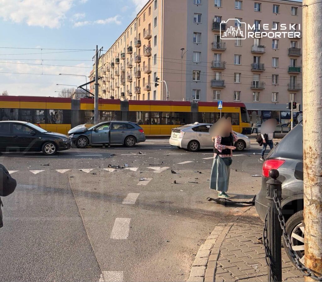 Na skrzyżowaniu z widocznymi uszkodzonymi pojazdami, kobieta w jasnej bluzie i długiej spódnicy stoi obok zniszczeń po wypadku.