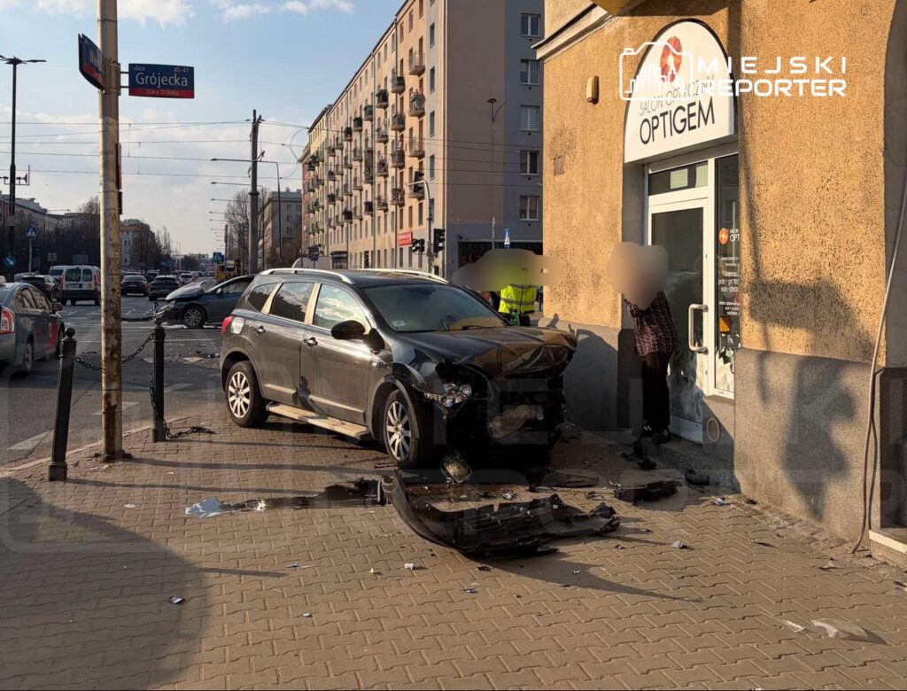 Czarny samochód osobowy z uszkodzonym przodem stoi na chodniku przed sklepem, obok niego mężczyzna rozmawia z osobą w budynku.