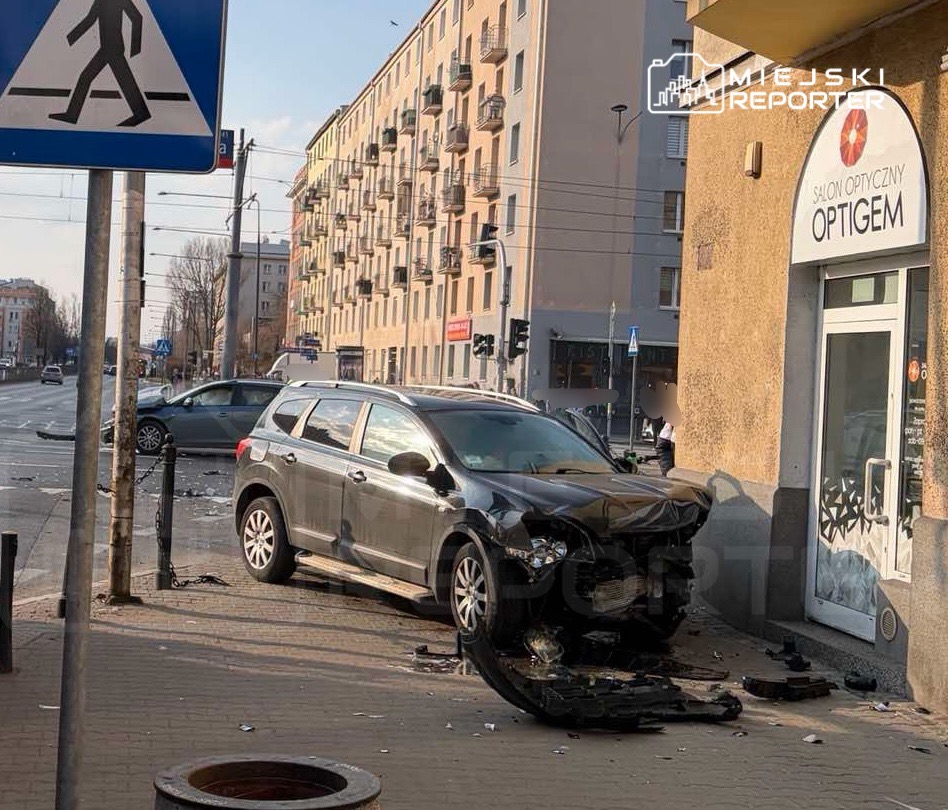 Czarny samochód osobowy z uszkodzoną przednią częścią stoi na chodniku obok salonu optycznego. W tle widoczna ulica z innymi pojazdami.