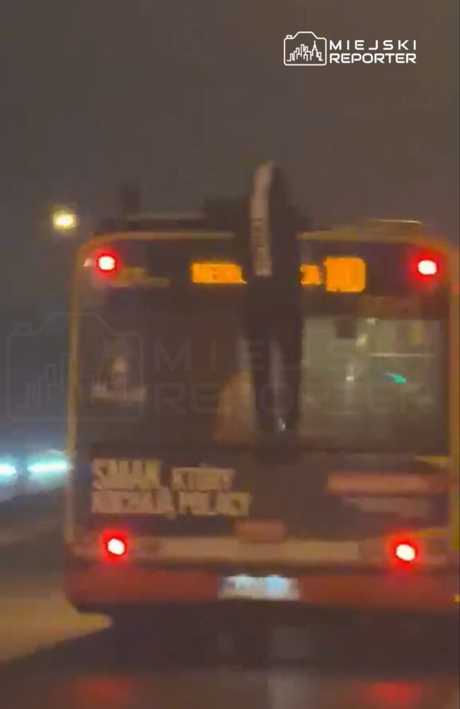 Mężczyzna wspinający się na tył autobusu w nocy, z widocznym wyświetlaczem kierunku „MEGA 100”.